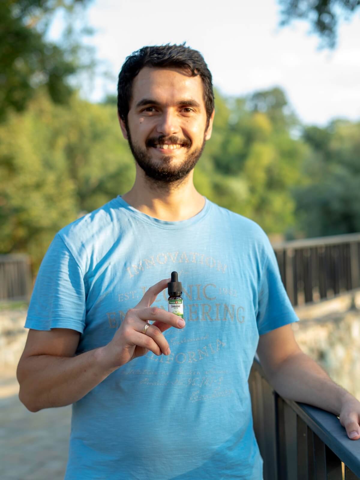 Smiling man in a blue T-shirt holding a bottle of Formula Swiss CBD oil in olive oil outdoors